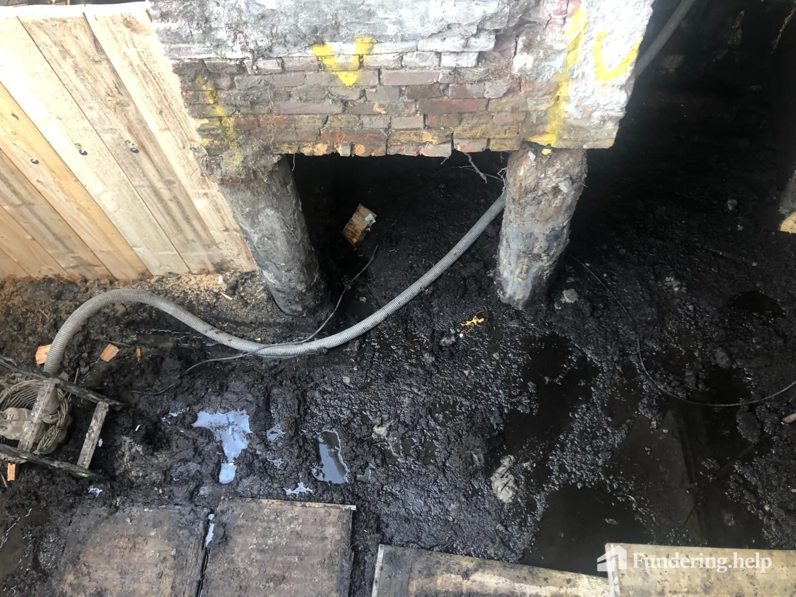 Houten paalkoppen in een ontgraven ruimte met een pompslang tijdens funderingsherstel
