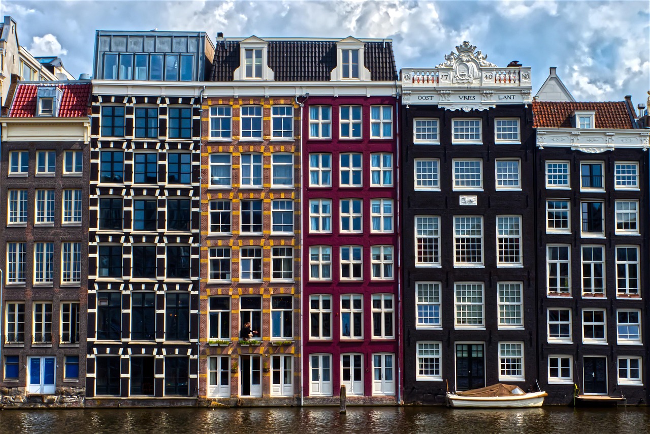 Rij gekleurde Amsterdamse grachtenpanden langs het water; de panden staan op houten palen in veen en klei en vertonen kenmerkende scheefstand door funderingszetting